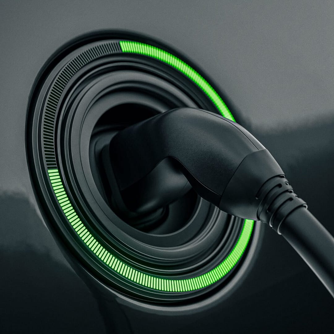 Close-up of an electric vehicle charging port with a plugged-in charger. A circular LED indicator surrounding the port glows green, indicating the charging status on a sleek, dark surface.