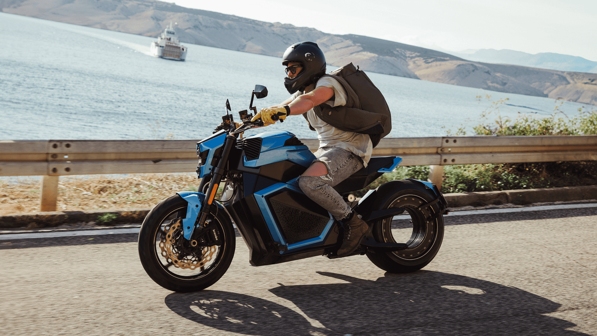A person wearing a helmet and backpack rides a futuristic blue motorcycle along a coastal road, with the ocean and a boat in the background. The scene is bright and sunny.
